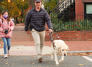 Hogan, a large yellow lab, guides Pat across the street on their way to the park. The guide dog team steps off the curb as a young photography student in a pink puffer jacket cradles her camera in the background. Pat sports a gray puffer jacket, khakis, and baseball cap.